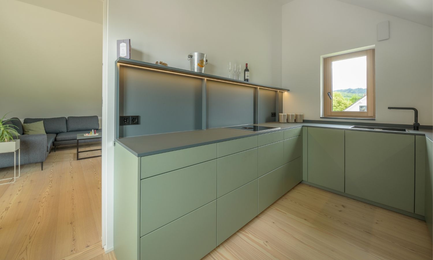 Cuisine moderne avec frontons verts mats, parquet en bois de larche, fenetre avec vue sur la verdure et salon attenant avec canape gris.