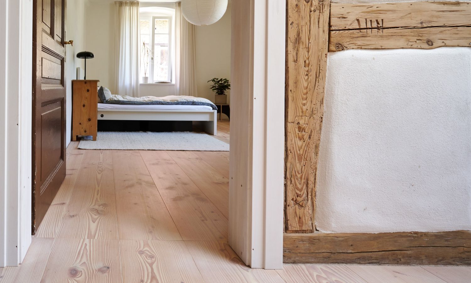 Blick durch einen Flur mit Douglasie Holzboden in ein Schlafzimmer mit Bett; Fenster und Vorhängen; rechts Wand mit sichtbaren Holzbalken