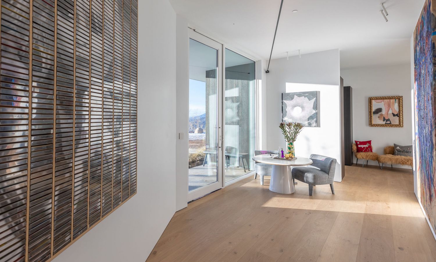 Piece lumineuse et moderne avec sol en bois de chene, grande oeuvre murale en lattes de bois a gauche, table ronde avec chaises et fleurs au centre, autre tableau sur le mur de droite et canape en arriere-plan.