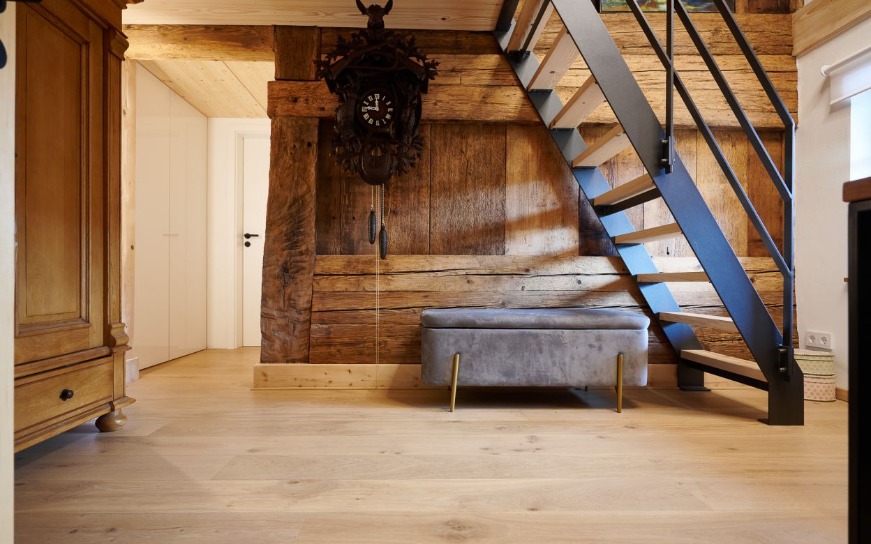 Holztreppe mit schwarzem Gelaender neben einer grauen Sitzbank vor einer Holzwand mit einer grossen Kuckucksuhr, links ein Holzmoebel und eine weisse Tuer.