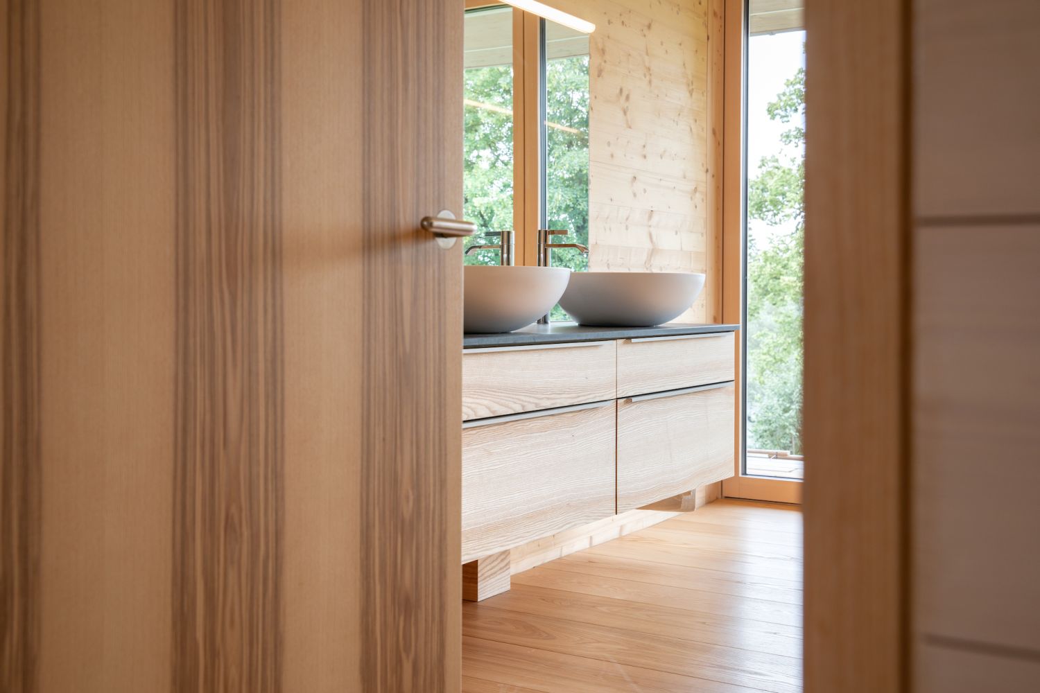 Offenes Holz Tuerblatt mit Griff, Blick in ein Badezimmer mit zwei weissen runden Waschbecken auf einem hellen Holz Waschtisch vor grossen Fenstern.
