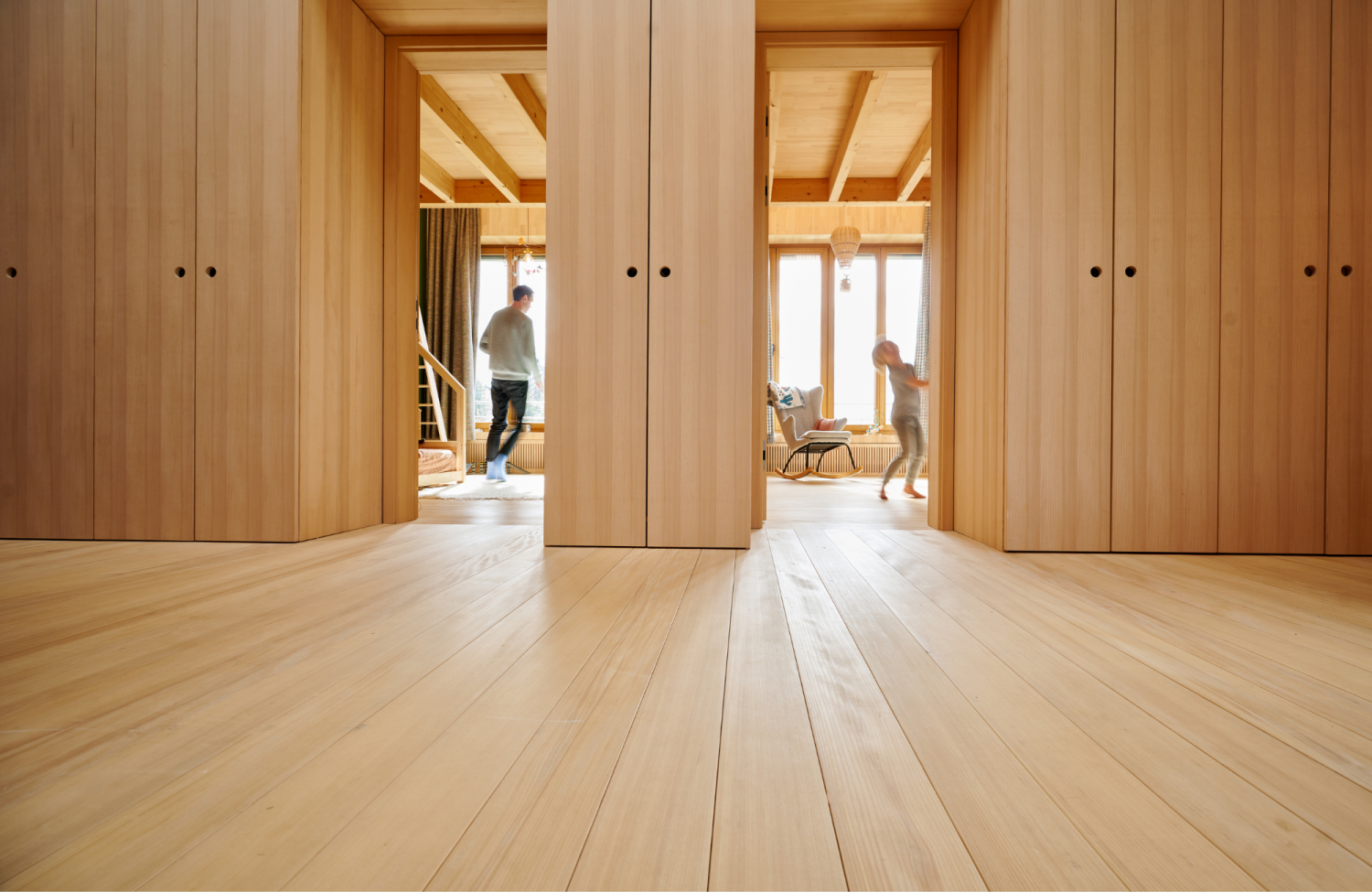 Blick auf hellen Holzboden und zwei offene Tueren mit Holzvertafelung, zwei Personen in Bewegung im Hintergrund.