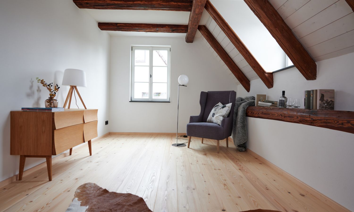 Dachzimmer mit sichtbaren Holzbalken, Weisstanne Holzboden, dunkelgrauem Sessel mit Kissen, Stehlampe und Sideboard mit Lampe und Dekoration.