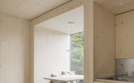 Moderner Innenraum mit hellen Holzflaechen, integrierter Sitzbank mit grauen Polstern und kleinem Esstisch vor Fenster mit Blick ins Grueene.