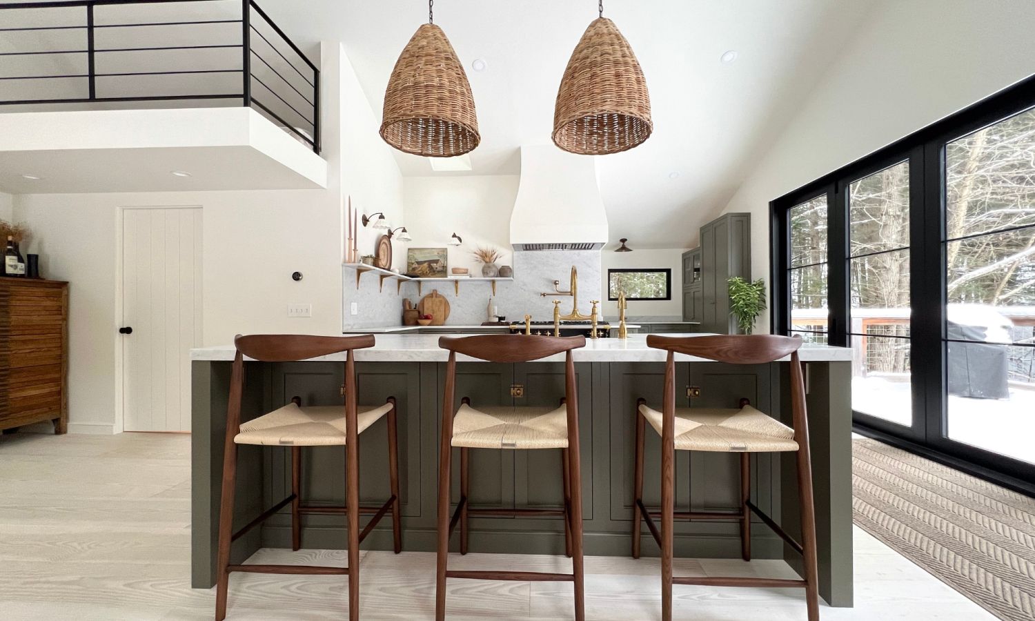 Modern kitchen with kitchen island, three wooden chairs with light seats, and two woven pendant lights above the island, large windows with views of a snowy landscape.