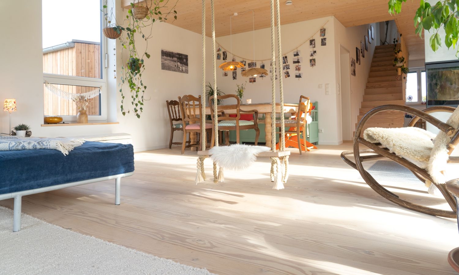 Espace de vie lumineux avec sol en bois, balançoire avec housse en fourrure blanche au centre, table à manger avec chaises à l’arrière, escalier à droite et plantes suspendues au plafond, avec parquet en douglas.