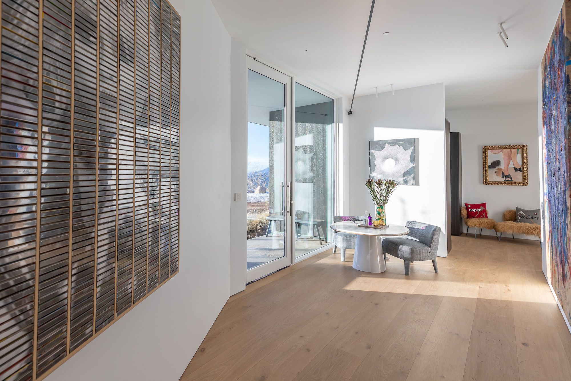 Moderner heller Raum mit Eiche Holzboden, grossem Wandbild aus Holzlatten links, rundem Tisch mit Stuehlen und Blumen in der Mitte, weiterem Bild an der Wand rechts und Sofa im Hintergrund.