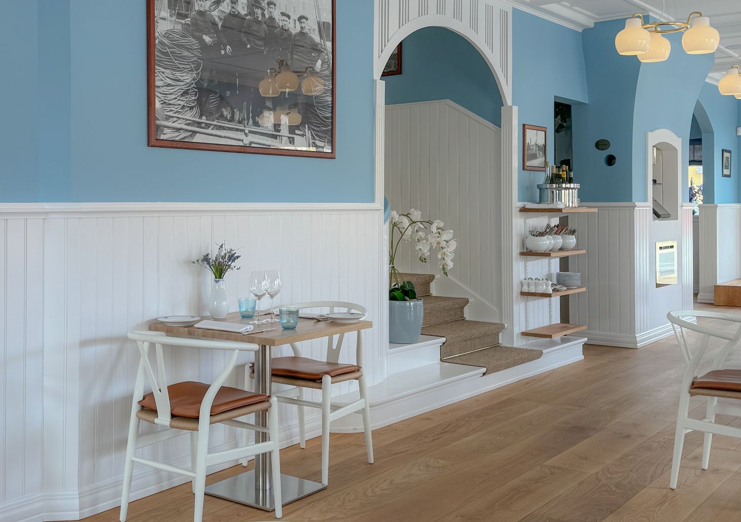 Hellblauer Gastraum mit weisser Holzvertafelung, Eiche Holzboden, zwei Tischen mit weissen Stuehlen und braunen Sitzkissen, Treppe und Wandregal mit Geschirr.