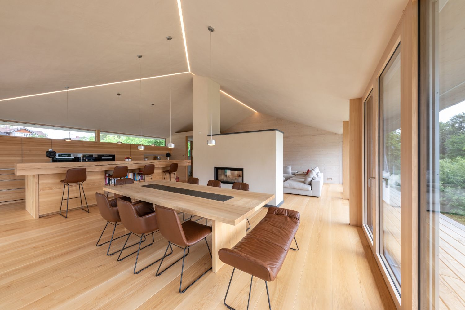 Moderner Essbereich mit langem Holztisch, braunen Lederstuehlen, offener Kueche und grossem Fenster mit Blick ins Gruene.