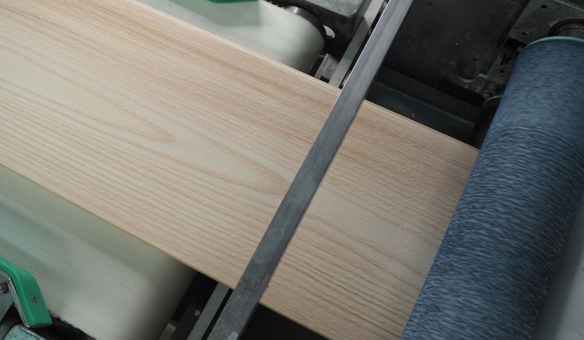 Wooden board with light brown grain lies on a machine next to a roll of dark blue fabric.