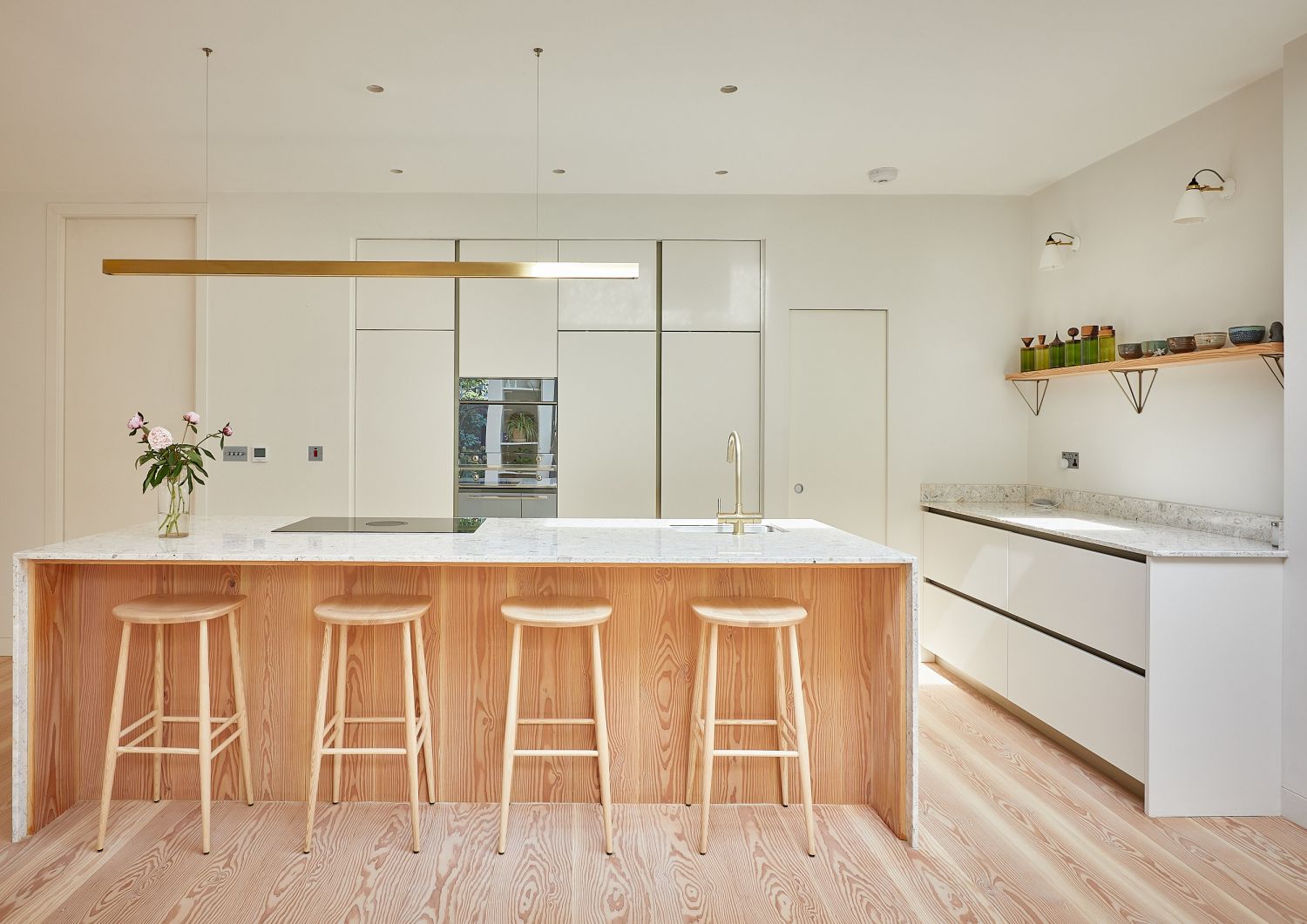 Cuisine moderne avec sol en bois de douglas, îlot avec quatre tabourets en bois, façades blanches et longue lampe de plafond étroite.