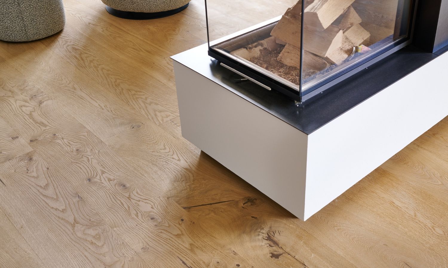 Modern rectangular fireplace with glass front and firewood on oak flooring, two round stools in the background.