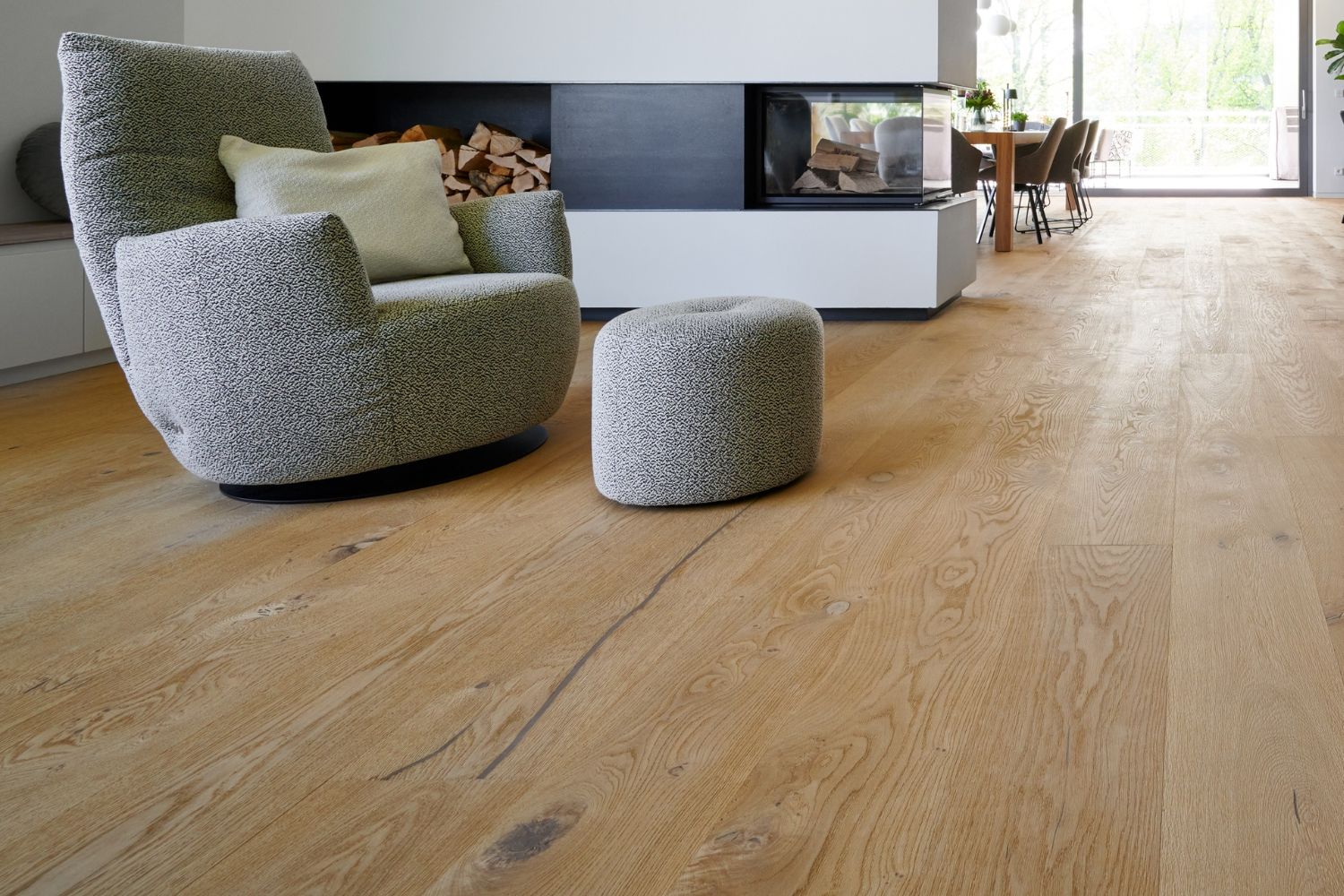 Fauteuil rembourre gris clair avec pouf assorti sur un sol en chene devant une cheminee moderne avec rangement pour le bois.