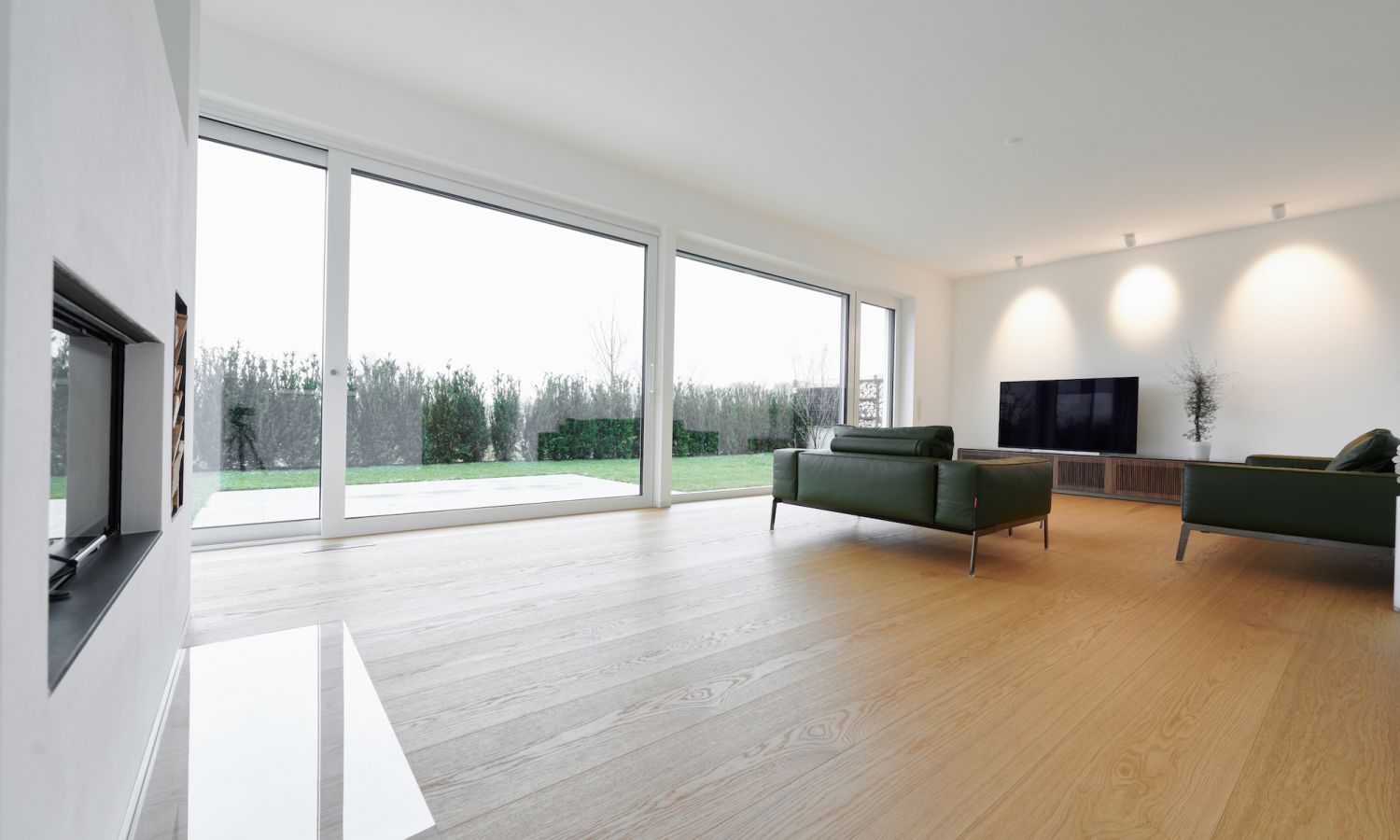 Salon lumineux avec grande baie vitree, deux canapes verts, televiseur a ecran plat sur mur blanc et sol en chene.