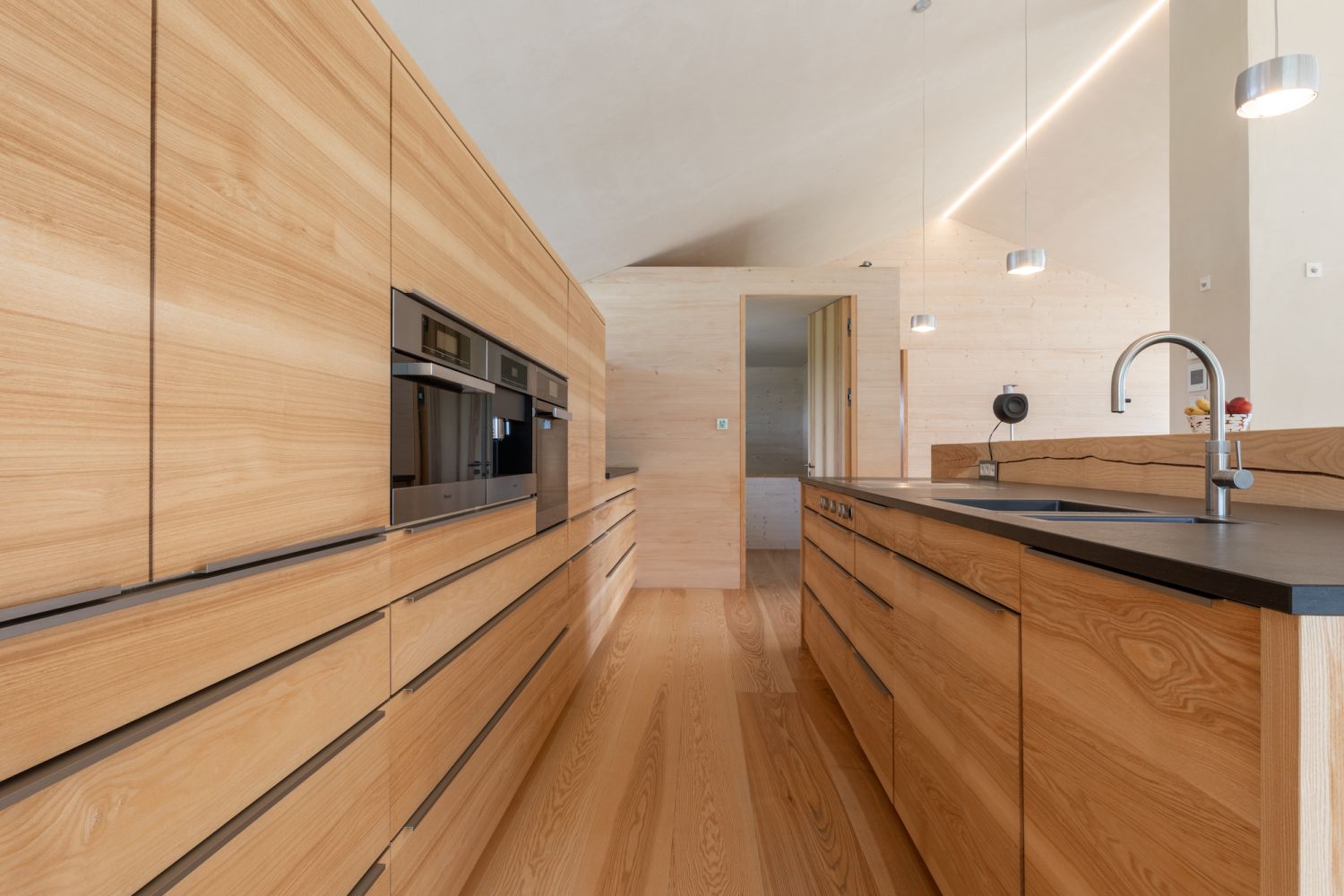 Modern kitchen with wooden fronts, built-in oven, kitchen island with sink and three hanging lights on ash wood flooring.