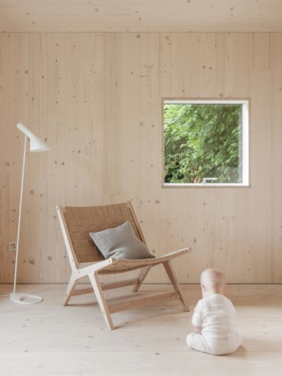 Kind in weissem Strampler sitzt auf Weisstanne Holzboden vor einem Holzstuhl mit grauem Kissen, daneben steht eine weisse Stehlampe, im Hintergrund ein Fenster mit Blick auf gruene Pflanzen.