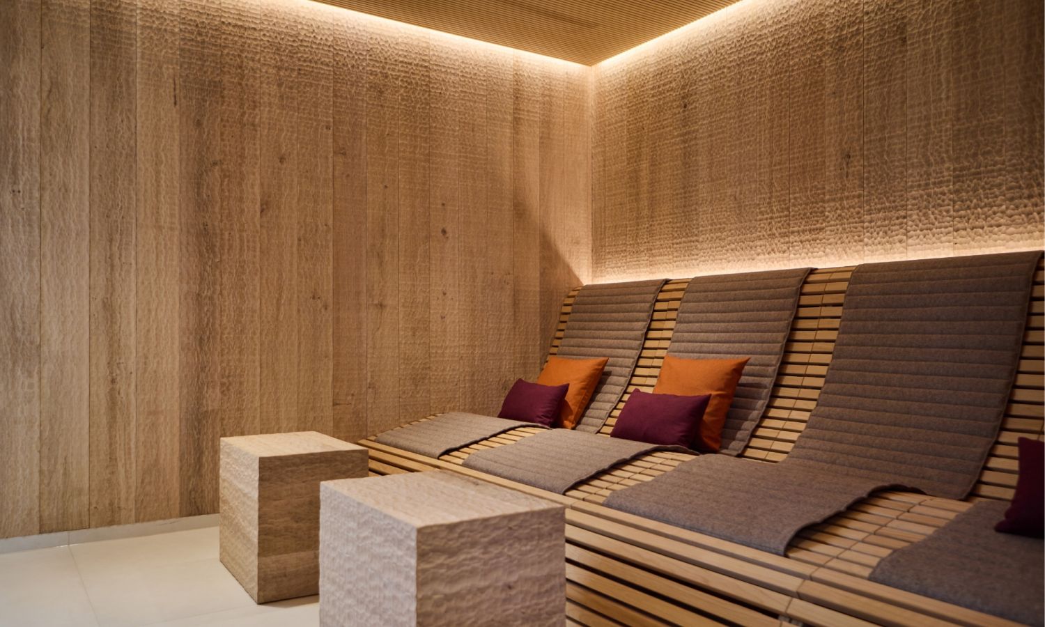 Three loungers and two square stools in a spa area. The walls are clad with wavy oak panels, and the loungers are adorned with red and orange cushions.