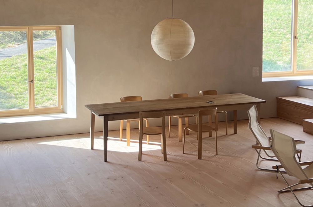 Une salle à manger dans une ferme rénovée avec de larges lames en sapin Douglas. Elle est équipée d’une table en bois simple, de chaises en bois modernes et d’une grande lampe ronde blanche.