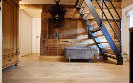 Holztreppe mit schwarzem Gelaender neben einer grauen Sitzbank vor einer Holzwand mit einer grossen Kuckucksuhr, links ein Holzmoebel und eine weisse Tuer.