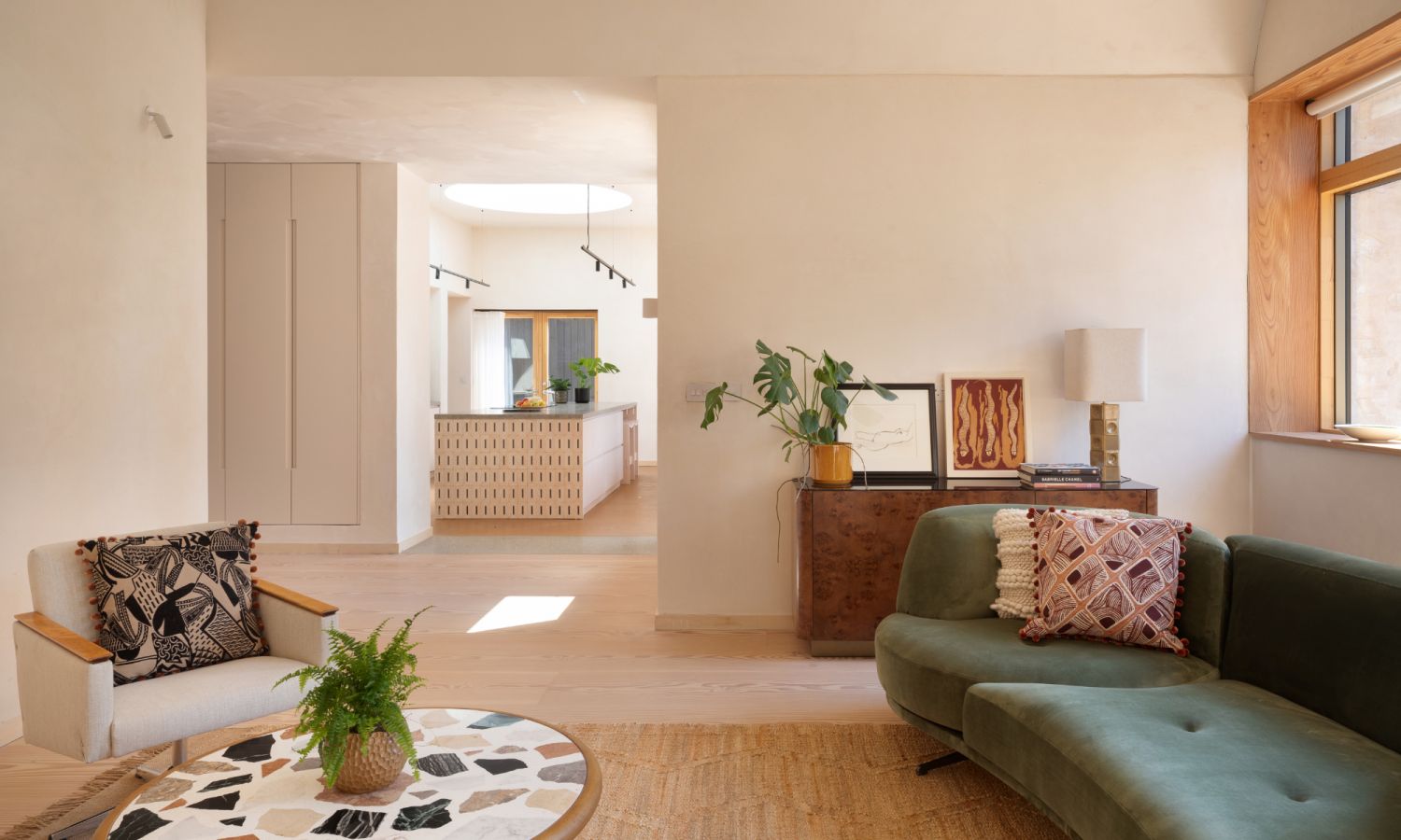 Un salon aux matériaux naturels. Tapis en fibres brun clair, table avec plateau en terrazzo, canapé en velours vert foncé et parquet clair en sapin Douglas. À l’arrière, la vue s’ouvre sur la cuisine.
