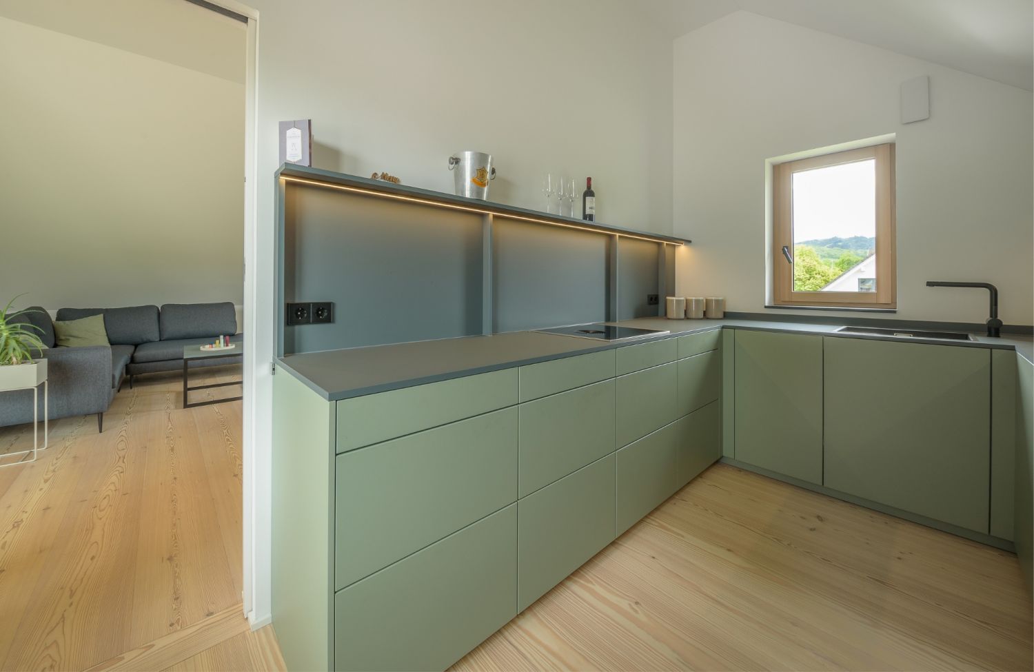 Cuisine moderne avec frontons verts mats, parquet en bois de larche, fenetre avec vue sur la verdure et salon attenant avec canape gris.