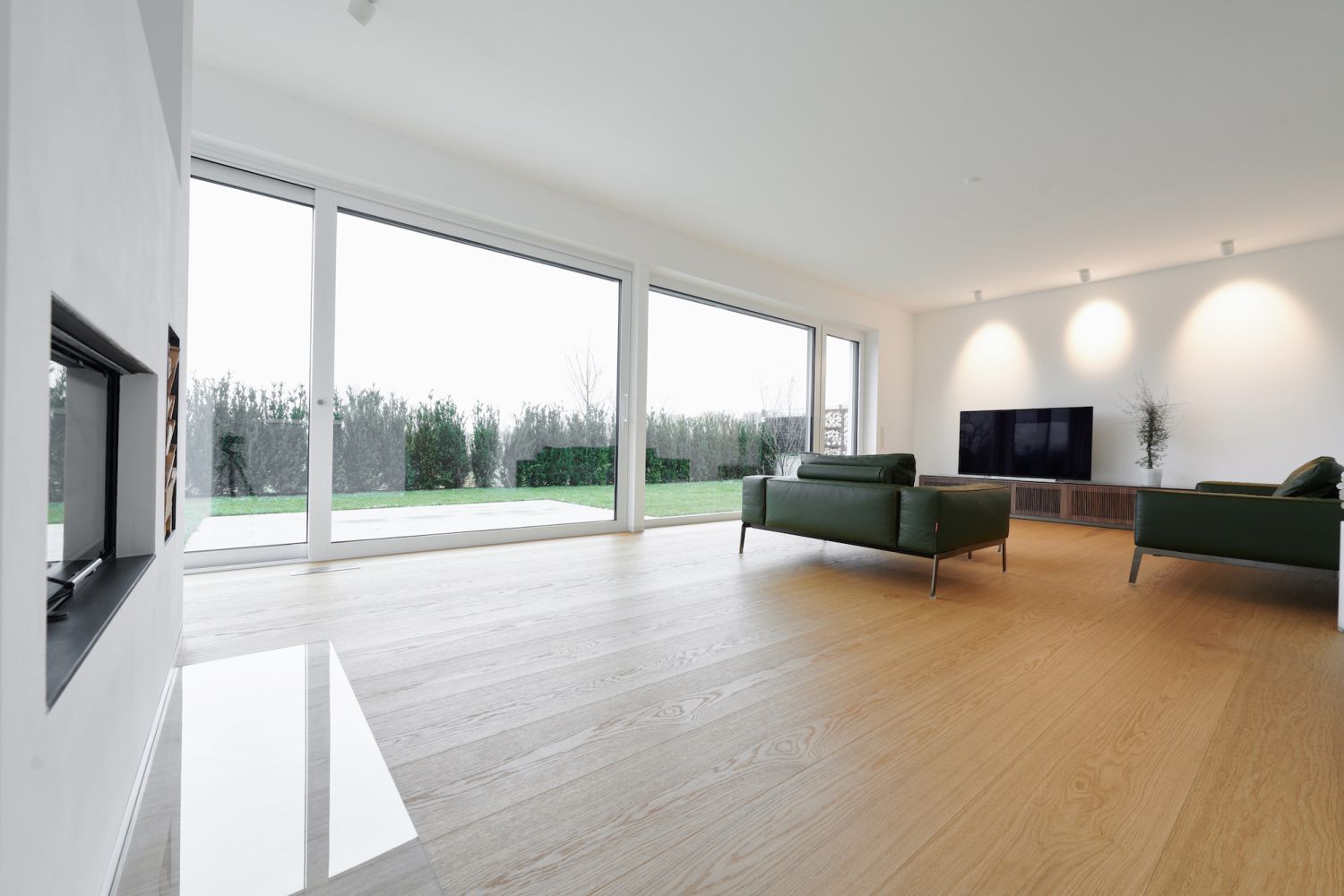 Salon lumineux avec grande baie vitree, deux canapes verts, televiseur a ecran plat sur mur blanc et sol en chene.