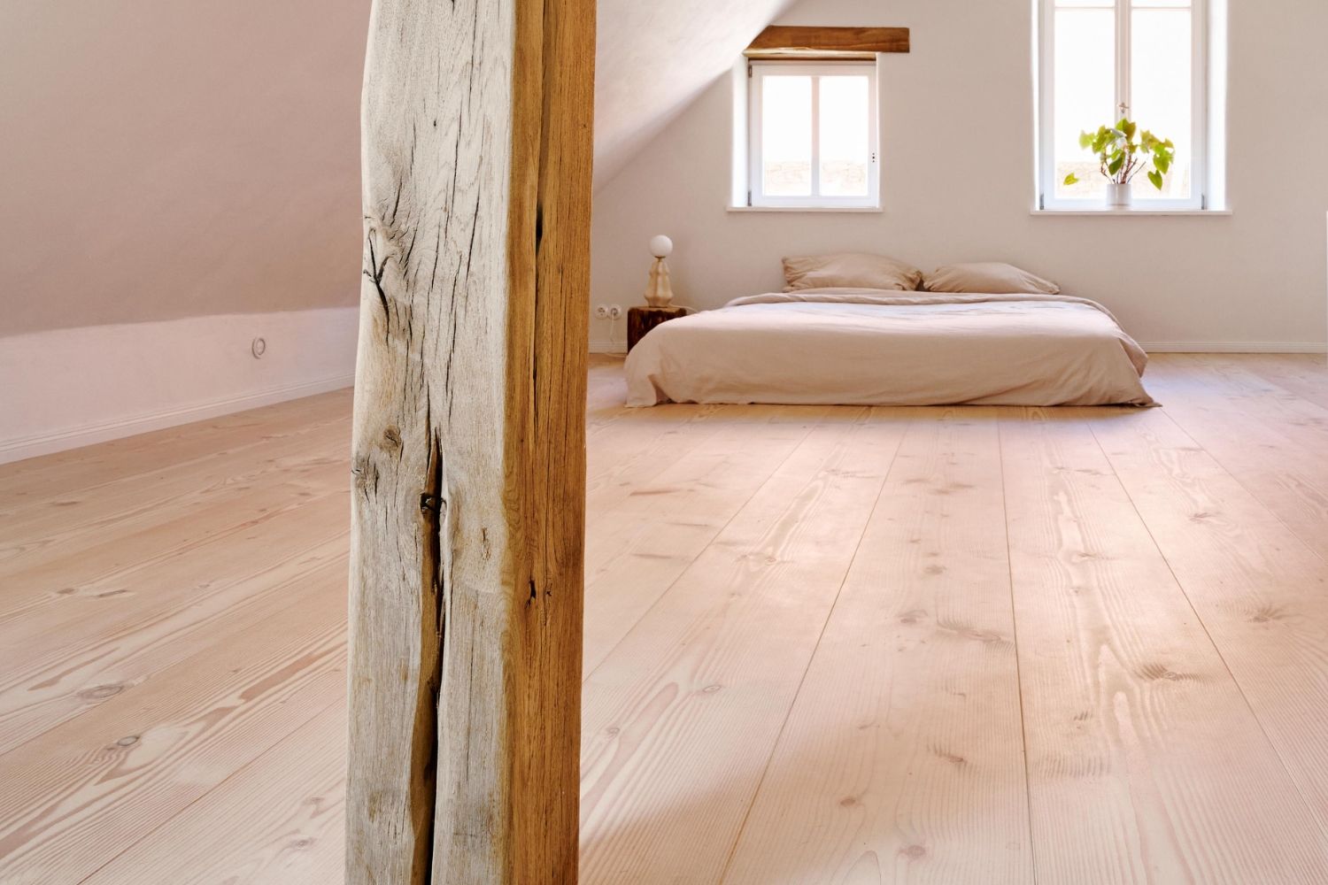 Schlafzimmer mit Holzbalken; Douglasie Holzboden; ein Bett vor zwei Fenstern; Pflanzen auf der Fensterbank