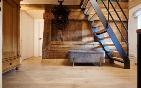 Holztreppe mit schwarzem Gelaender neben einer grauen Sitzbank vor einer Holzwand mit einer grossen Kuckucksuhr, links ein Holzmoebel und eine weisse Tuer.