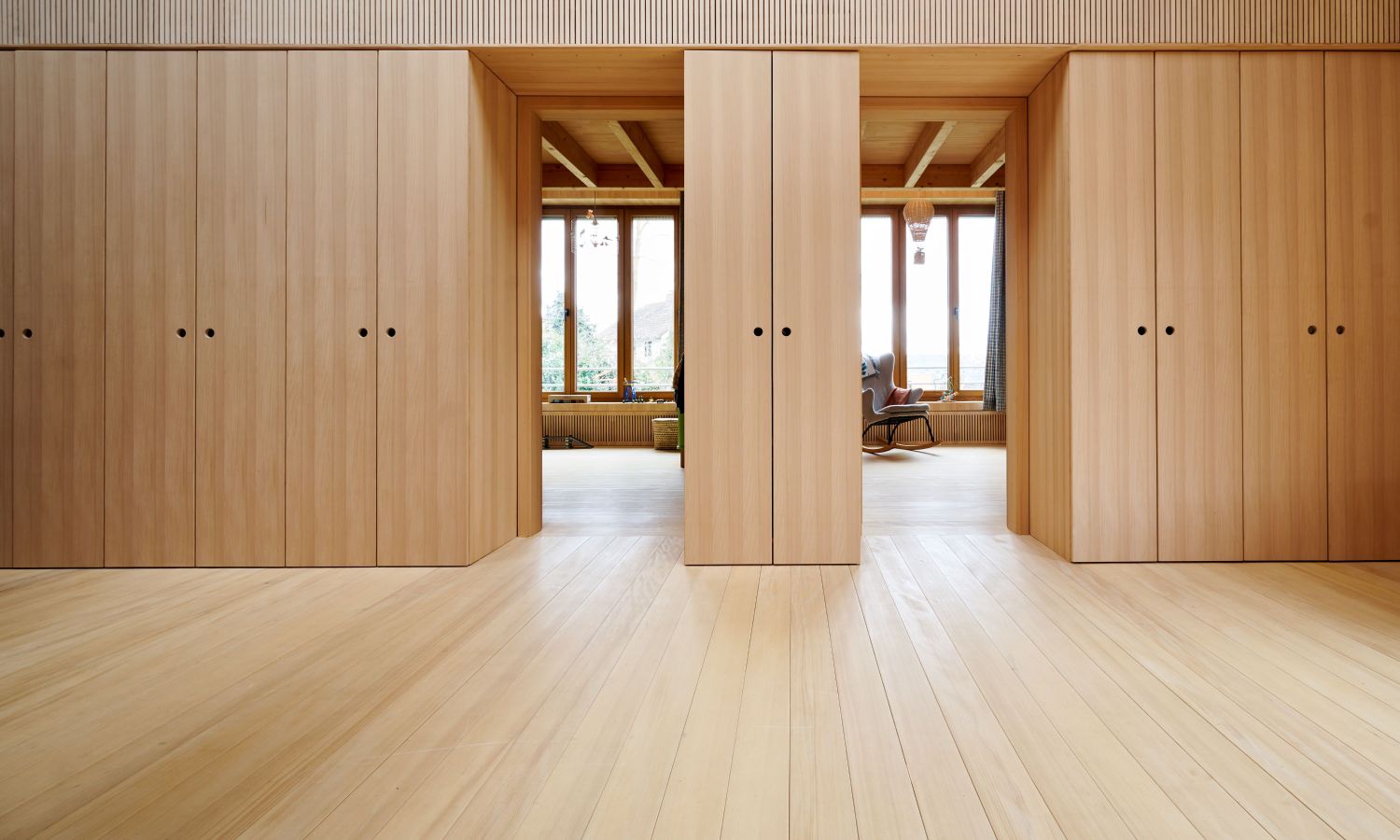Grande piece avec des sols en sapin blanc et plusieurs grands placards en bois simples avec poignées noires, menant a un coin salon avec de grandes fenetres.