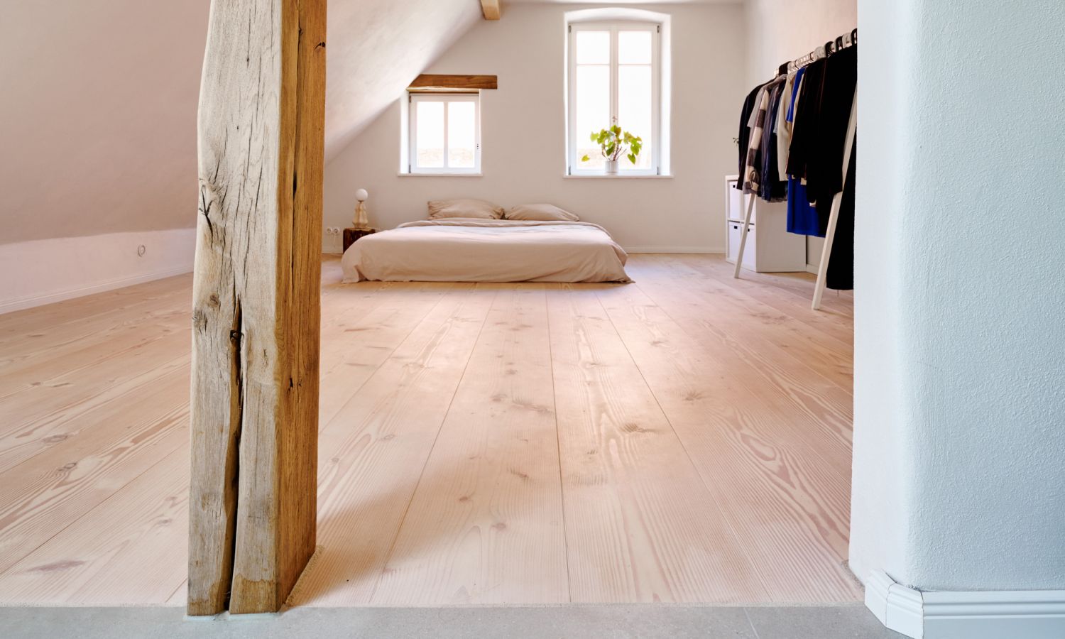Schlafzimmer mit Holzbalken; großem Fenster; niedrigem Bett; offenem Kleiderständer mit mehreren Kleidungsstücken