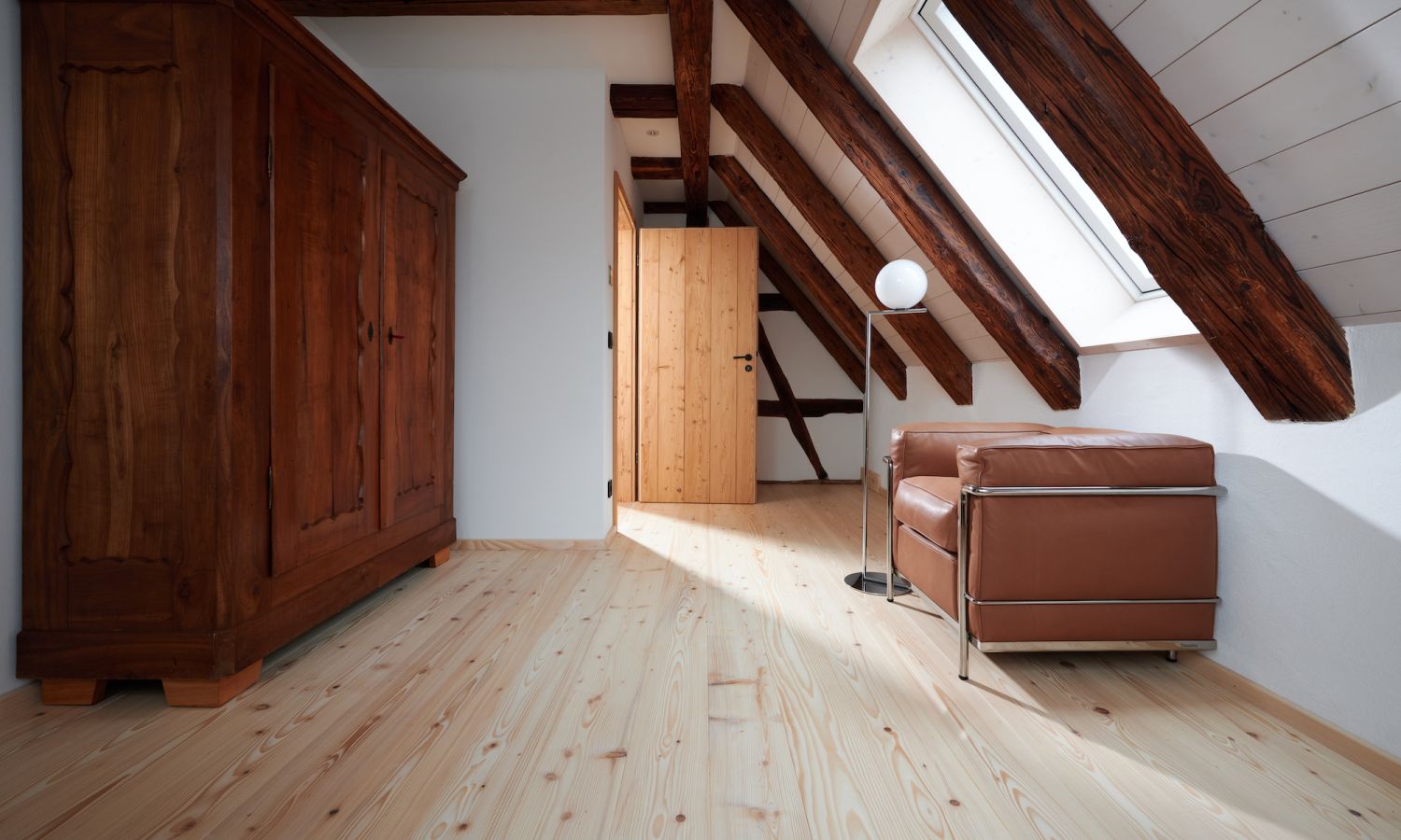 Dachzimmer mit sichtbaren Holzbalken, braunem Ledersessel, Stehlampe und großem Holzschrank auf hellem Holzboden.