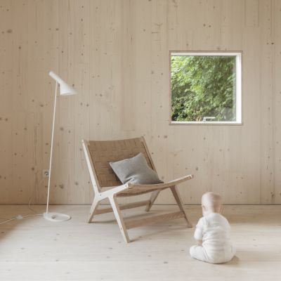 Enfant en grenouillere blanche assis sur un sol en bois clair devant une chaise en bois avec coussin gris, a cote d’une lampe sur pied blanche et d’une fenetre avec vue sur la verdure.