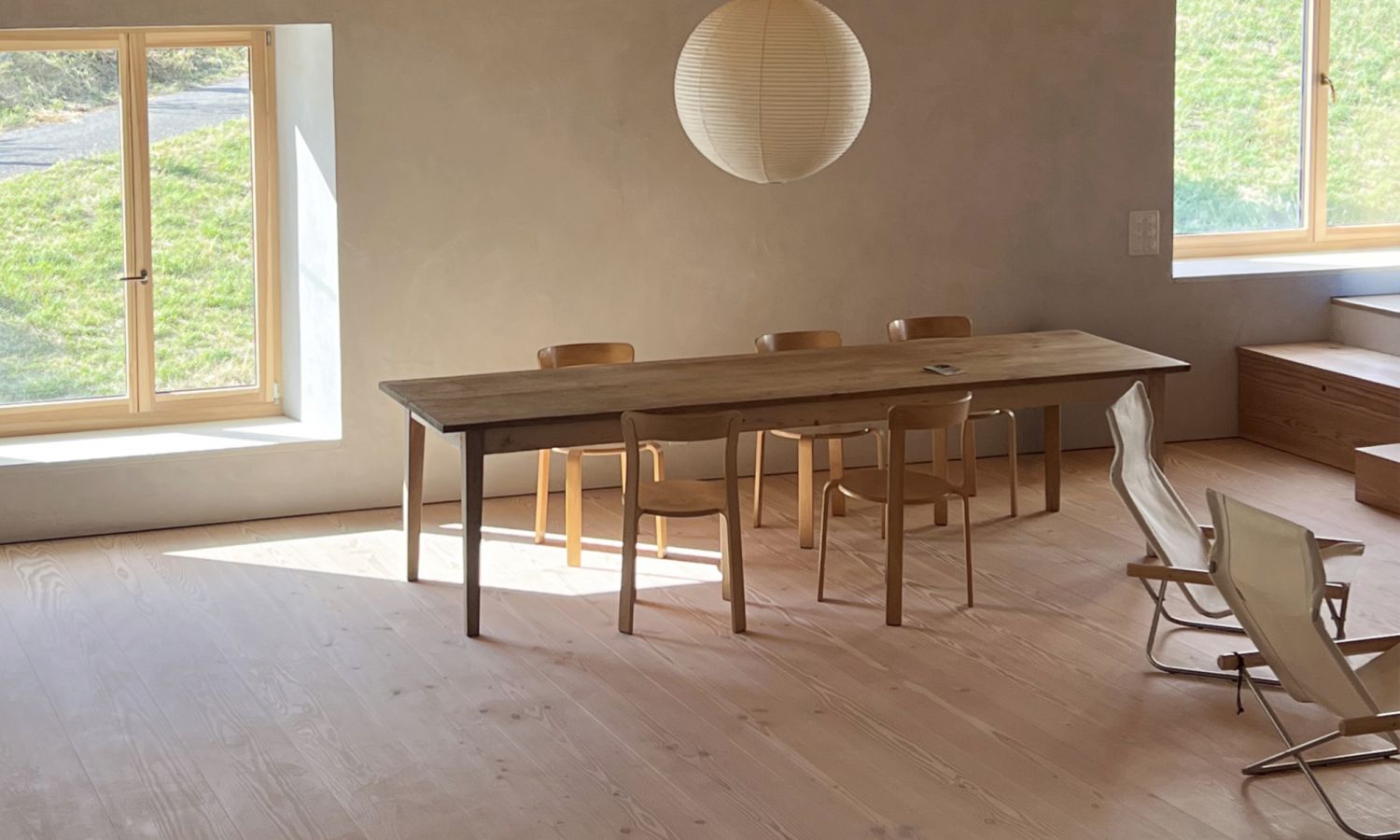 Une salle à manger dans une ferme rénovée avec de larges lames en sapin Douglas. Elle est équipée d’une table en bois simple, de chaises en bois modernes et d’une grande lampe ronde blanche.