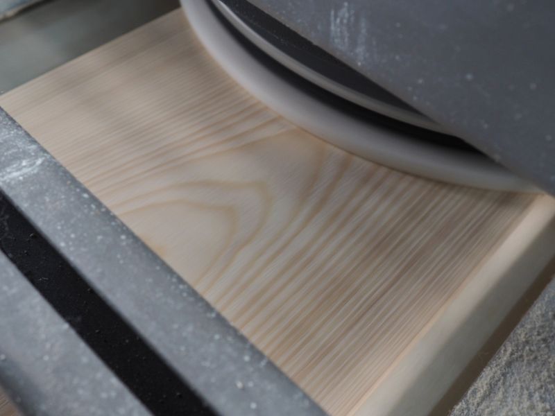 Wooden board being processed by a sanding machine, visible grain and dust particles on the surface.