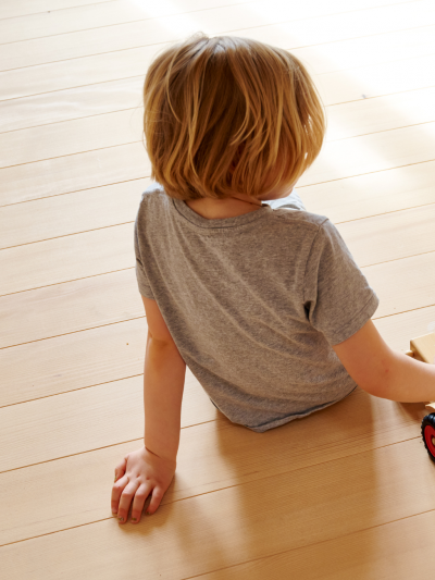 Kind mit kurzem, blondem Haar sitzt auf Weisstanne Holzboden und haelt ein Holzspielzeugauto mit roten Raedern.