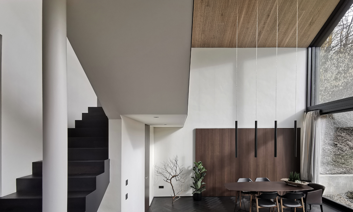 Moderner Essbereich mit dunklem Fischgraetparkett, Holztisch, sechs Stuehlen, haengenden schwarzen Pendelleuchten und grosser Fensterfront neben schwarzer Treppe