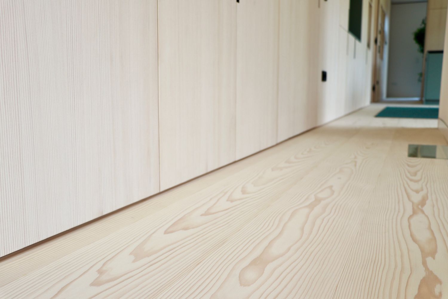 Vue sur un parquet en sapin blanc avec veinures visibles et un mur avec des portes d’armoires claires dans un couloir.