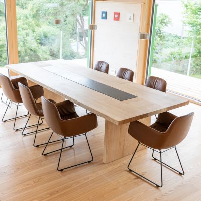 Salle a manger moderne avec longue table en bois clair et huit chaises marron avec pieds en metal dans une piece avec grandes fenetres et vue sur la verdure.