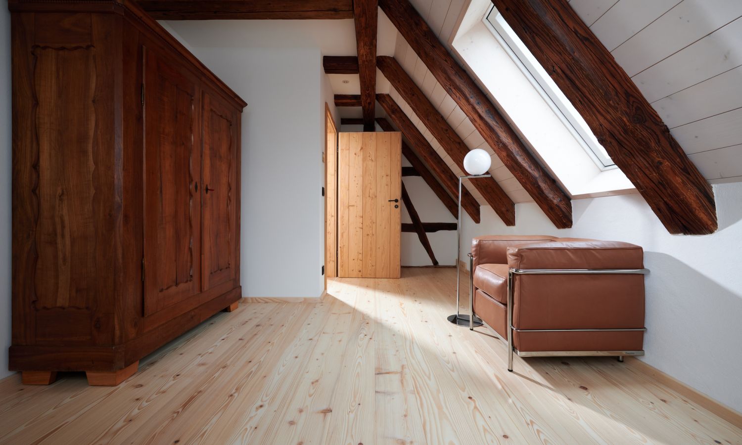 Dachzimmer mit sichtbaren Holzbalken, braunem Ledersessel, Stehlampe und großem Holzschrank auf hellem Holzboden.