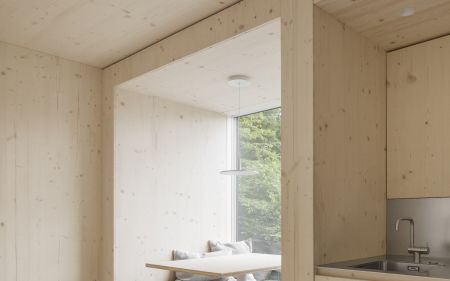 Moderner Innenraum mit hellen Holzflaechen, integrierter Sitzbank mit grauen Polstern und kleinem Esstisch vor Fenster mit Blick ins Grueene.