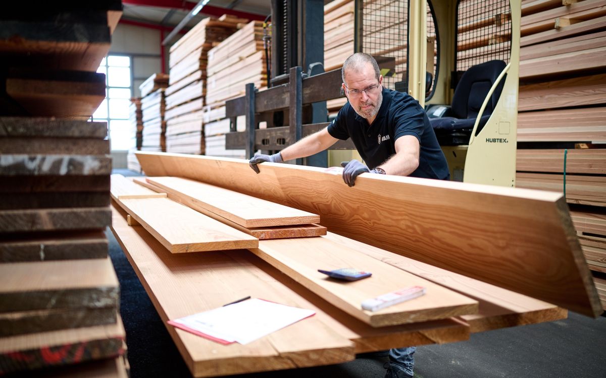 Un homme portant des gants souleve de longues planches en bois claires dans un entrepot avec des rayonnages et un chariot elevateur en arriere-plan.