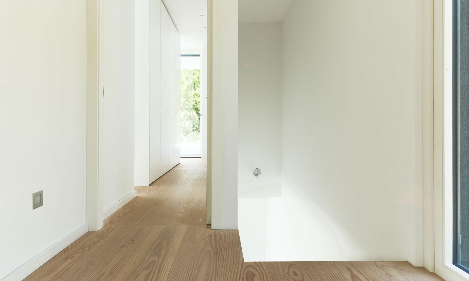 Couloir minimaliste avec sol en bois de douglas et murs blancs; à droite, une fenêtre partiellement visible; un luminaire encastré sur le mur de l’escalier éclaire vers le bas