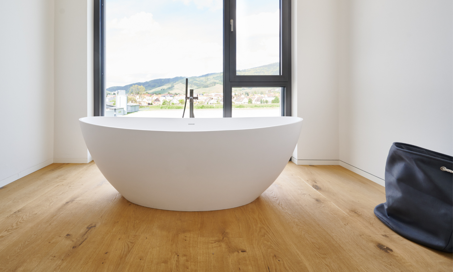 Baignoire blanche autoportante devant une grande fenêtre avec vue sur les collines, plancher en chene, sac noir a droite.