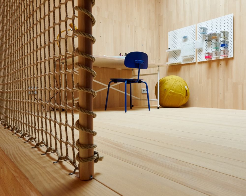 Piece avec panneaux en bois clair, chaise bleue, table blanche, pouf jaune et deux panneaux perforés blancs au mur avec de petits objets.