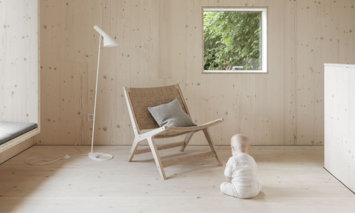 Enfant en grenouillere blanche assis sur un sol en bois clair devant une chaise en bois avec coussin gris, a cote d’une lampe sur pied blanche et d’une fenetre avec vue sur la verdure.