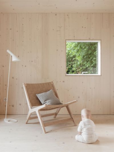 Kind in weissem Strampler sitzt auf Weisstanne Holzboden vor einem Holzstuhl mit grauem Kissen, daneben steht eine weisse Stehlampe, im Hintergrund ein Fenster mit Blick auf gruene Pflanzen.