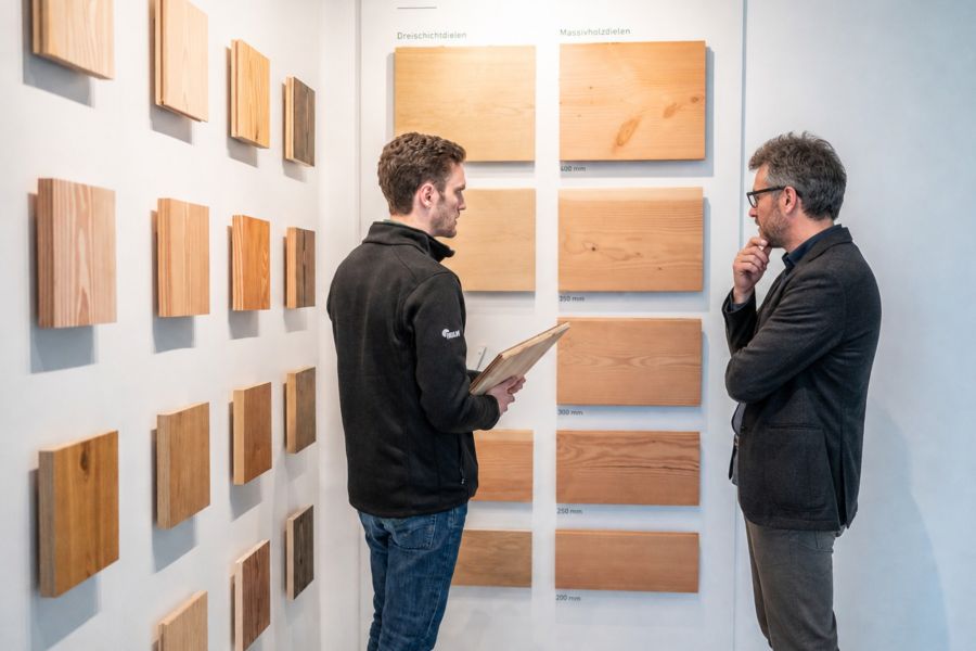 Zwei Männer stehen vor einer Wand mit Holzboden Mustern. Der linke Mann hat ein Muster in der Hand, der andere Mann hört aufmerksam zu.