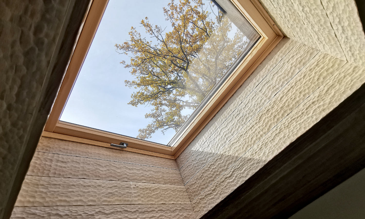 Blick durch ein rechteckiges Dachfenster mit Eiche Wandpaneele auf einen Baum mit gelblichen Blaettern vor blauem Himmel