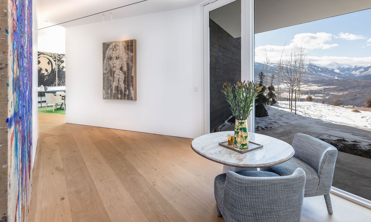 Modern living area with oak wood flooring, round marble coffee table, two grey armchairs and large window with mountain view.