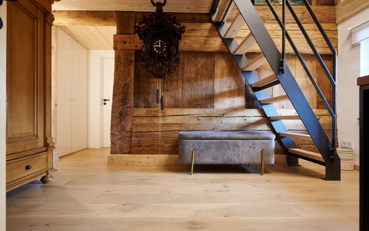 Holztreppe mit schwarzem Gelaender neben einer grauen Sitzbank vor einer Holzwand mit einer grossen Kuckucksuhr, links ein Holzmoebel und eine weisse Tuer.